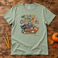 A sage green graphic t-shirt laid flat on a rustic wooden background, styled with two yellow pencils, a small orange pumpkin, and autumn leaves. The t-shirt features a colorful cartoon graphic of three cats stacked on books with the text 'MY CLASS IS FULL OF CHARACTERS'.