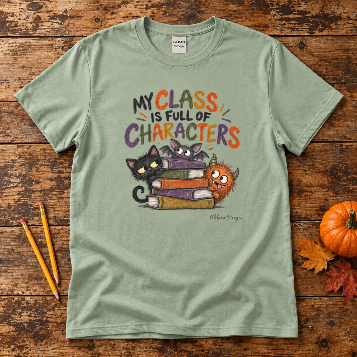 A sage green graphic t-shirt laid flat on a rustic wooden background, styled with two yellow pencils, a small orange pumpkin, and autumn leaves. The t-shirt features a colorful cartoon graphic of three cats stacked on books with the text 'MY CLASS IS FULL OF CHARACTERS'.