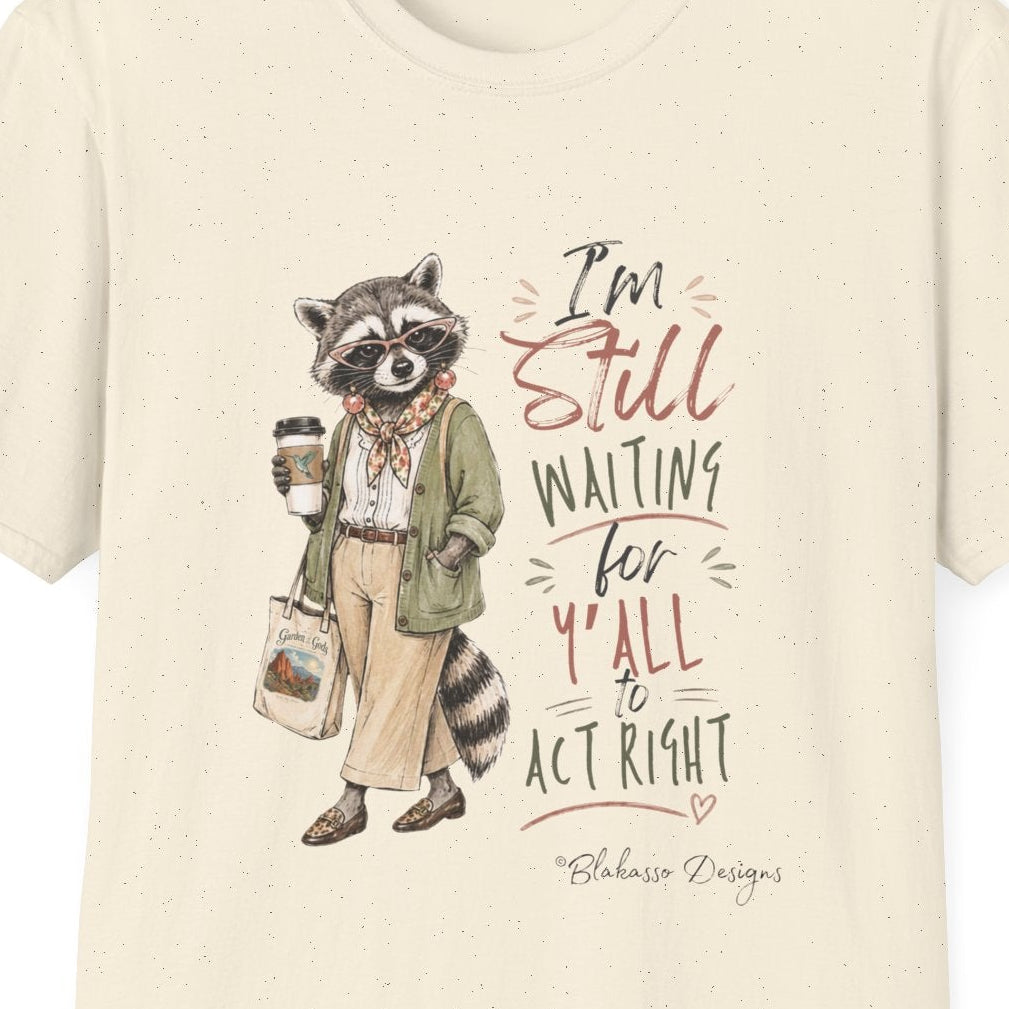 An off-white short-sleeve crewneck t-shirt featuring a graphic of a raccoon dressed in a green coat holding a coffee cup next to the text 'I'm Still WAITING for Y'all to ACT RIGHT' by designer Blakasso Designs.