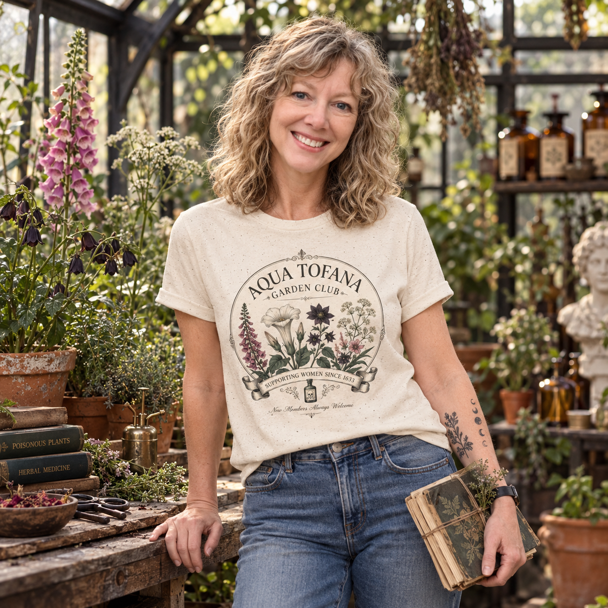 A smiling woman with curly blonde hair stands in a greenhouse wearing a cream graphic T-shirt that reads 'AQUA TOFANA GARDEN CLUB' with botanical illustrations, paired with blue jeans and holding a small book.