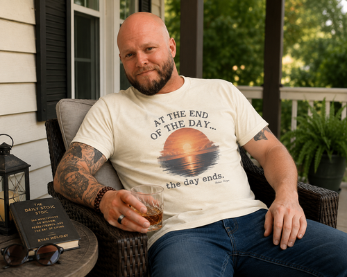Man relaxing on a porch wearing a Natural At the End of the Day dry humor sunset T-shirt from Blakasso Designs.