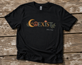 Black COEXIST unisex T-shirt displayed on a weathered wood surface with crescent moon, peace sign, lotus, and botanical artwork.