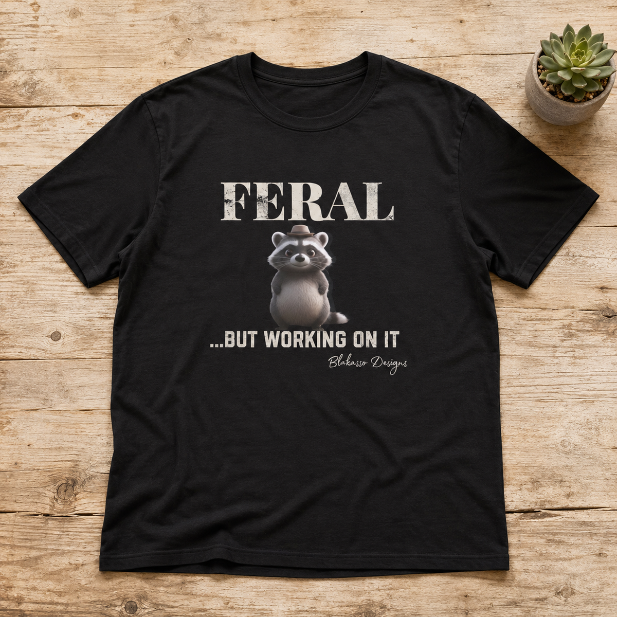 Black unisex Gildan Softstyle graphic tee featuring a small raccoon wearing a tiny hat beneath the phrase "Feral...But Working On It."