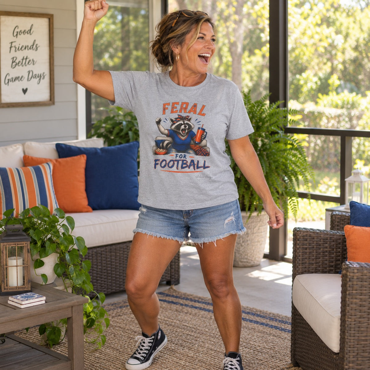 Woman wearing a gray Feral for Football raccoon shirt with denim shorts in an orange and blue game day setting.