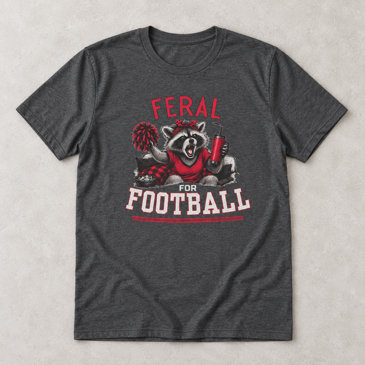 Dark heather Feral for Football Georgia T shirt flat lay featuring a cheering raccoon with a pom pom, drink tumbler, and bold red and white football graphic.