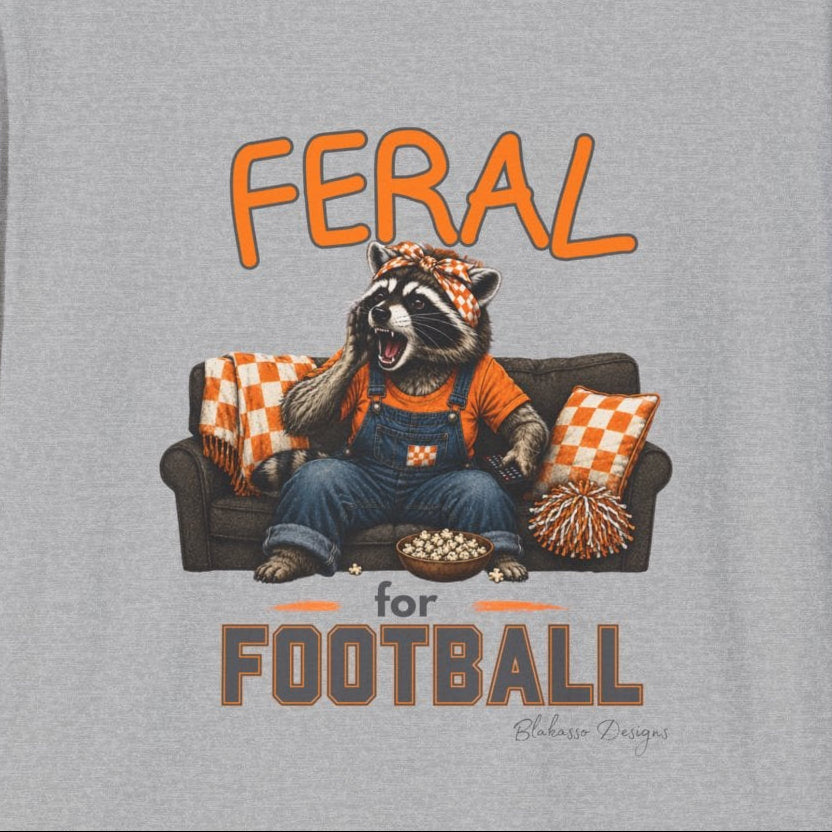 A heather gray short-sleeve crewneck t-shirt featuring a graphic of a raccoon sitting on a couch with a football, with the text 'FERAL for FOOTBALL' in orange and brown.