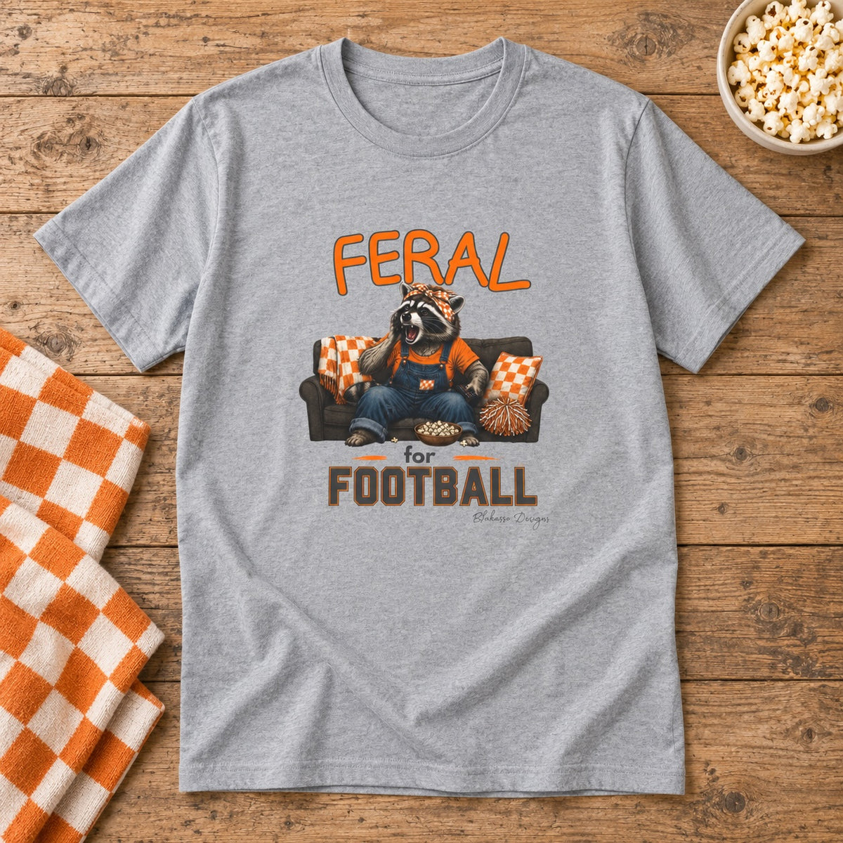 A heather gray short-sleeve t-shirt laid flat on a rustic wood surface, featuring a graphic of a raccoon  sitting on a couch with the words 'FERAL for FOOTBALL' in orange and brown. An orange-and-white checkered towel, a bowl of popcorn, and a wooden tabletop are visible around the shirt.