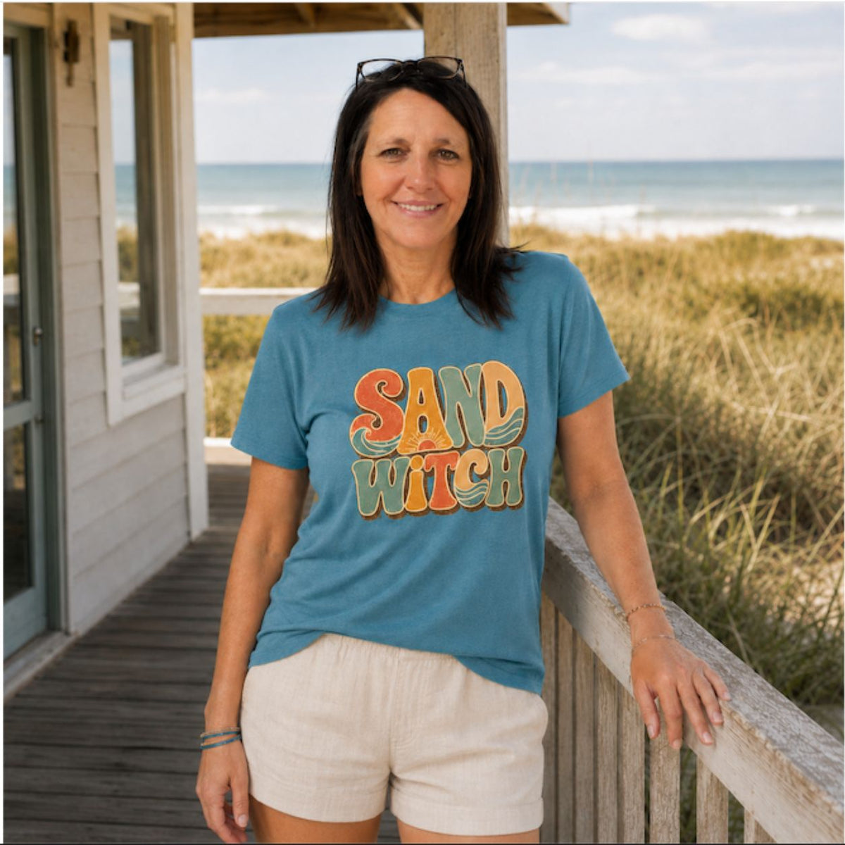 A woman stands on a beach house porch wearing a blue graphic t-shirt that reads 'SAND WITCH' in a retro orange and yellow font, paired with cream shorts. The background shows a wooden railing, beach grass, and the ocean.