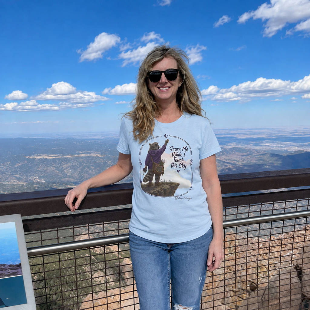 Wman wearing a light blue Scuse Me While I Touch This Sky raccoon graphic T-shirt in a relaxed outdoor setting.
