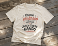 Natural unisex T-shirt laid on a weathered porch surface featuring the Choose Kindness Always or Catch These Hands text design.