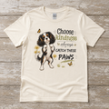 Natural Gildan Softstyle t-shirt laid flat on a weathered wood surface featuring a tricolor Cavalier dog design with the phrase Choose Kindness Always or Catch These Paws.