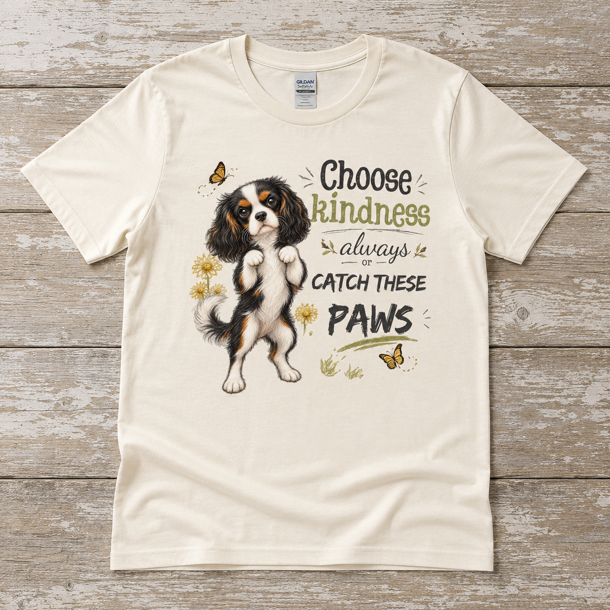 Natural Gildan Softstyle t-shirt laid flat on a weathered wood surface featuring a tricolor Cavalier dog design with the phrase Choose Kindness Always or Catch These Paws.