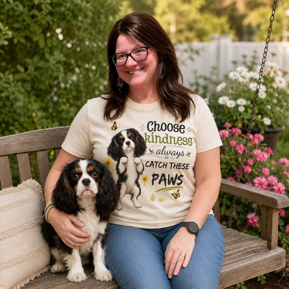 Woman wearing a natural t-shirt with a tricolor Cavalier dog design while sitting on a wooden glider in a flower-filled yard with Ref beside her.