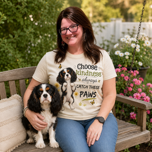 Woman wearing a natural t-shirt with a tricolor Cavalier dog design while sitting on a wooden glider in a flower-filled yard with Ref beside her.