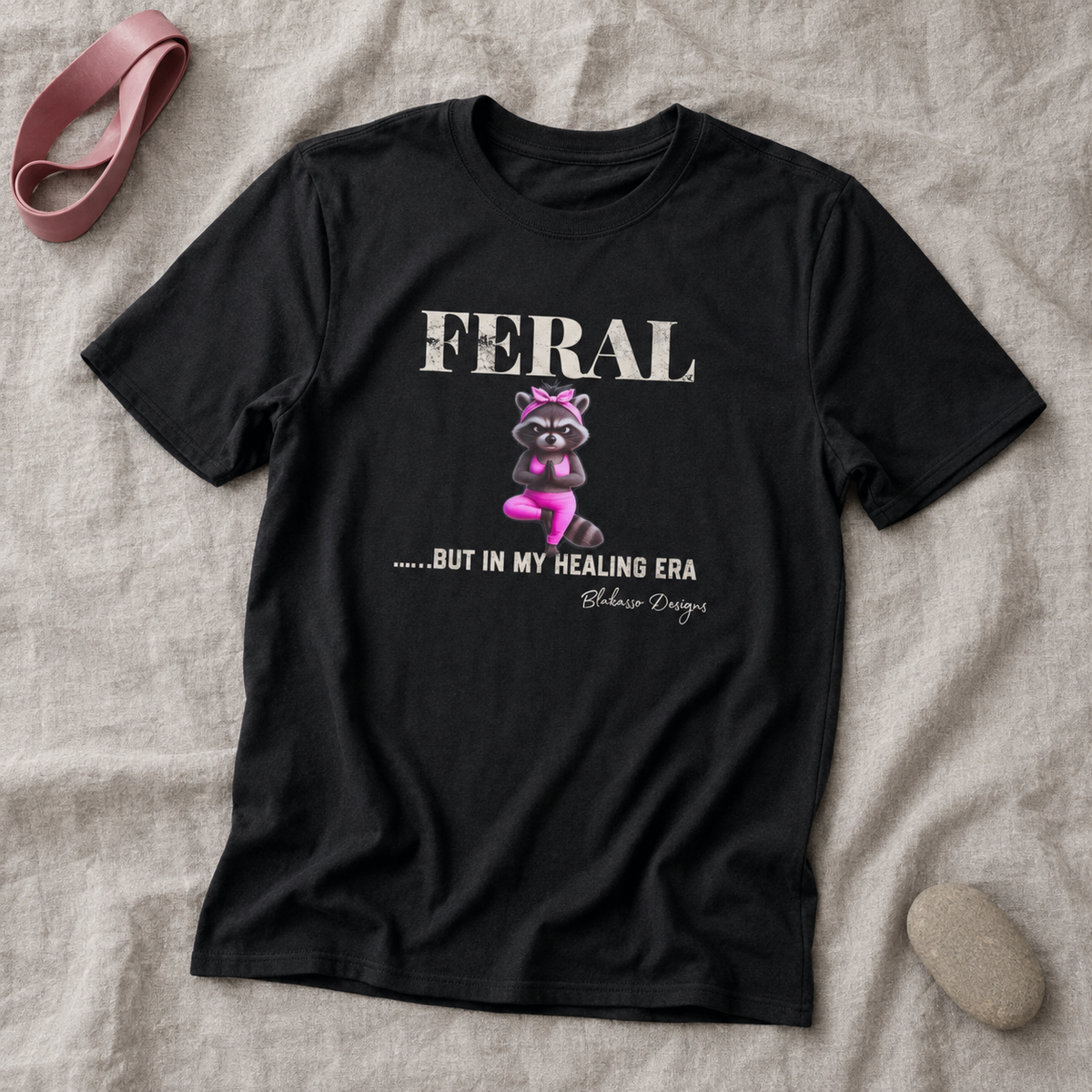 Black unisex graphic tee featuring a raccoon in pink yoga clothes beneath the words “Feral...But In My Healing Era.”