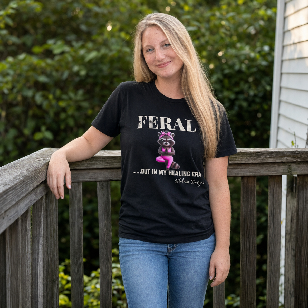 Woman wearing the black “Feral...But In My Healing Era” raccoon graphic tee outdoors on a wooden deck.
