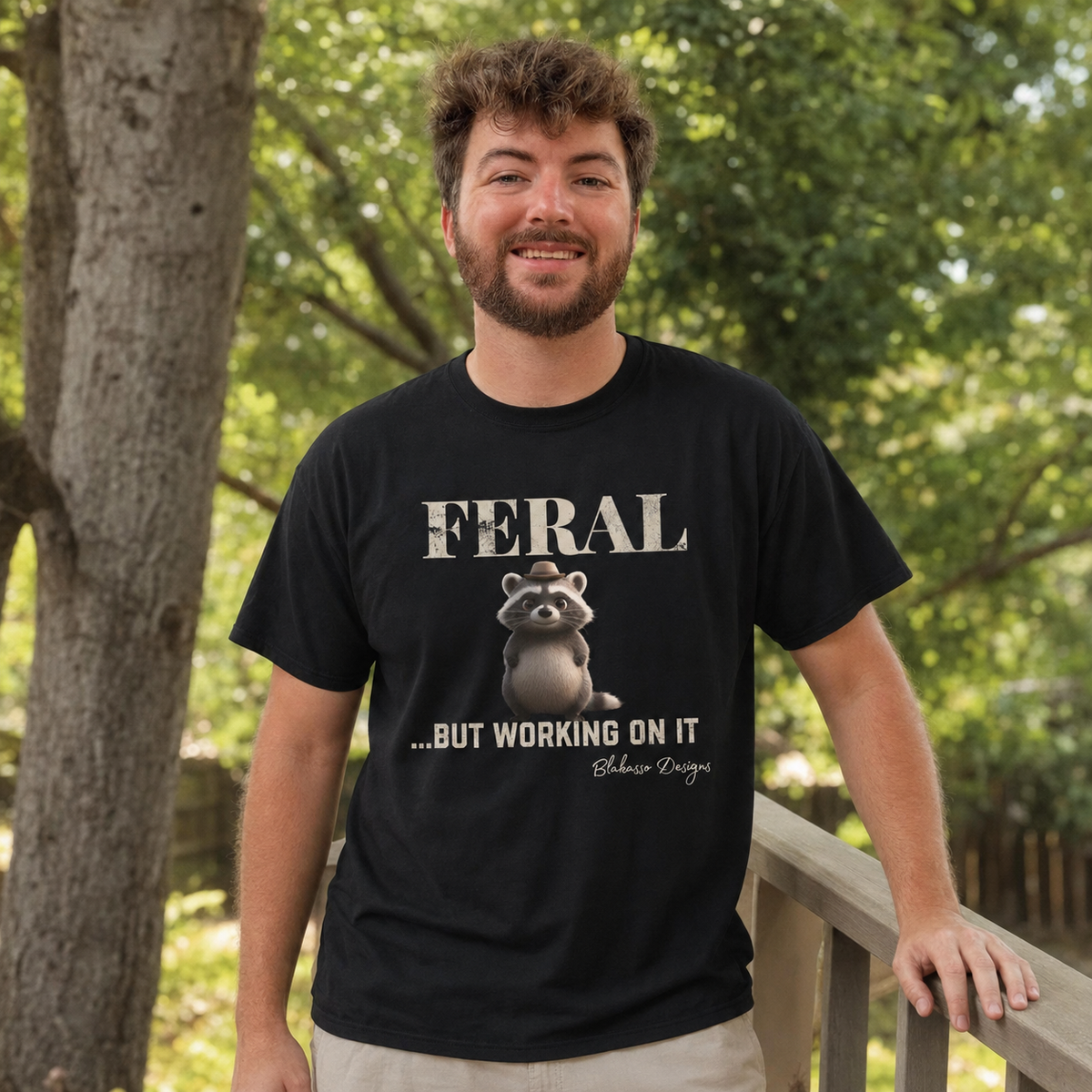 Young man wearing the "Feral...But Working On It" raccoon graphic tee while standing outdoors on a wooden deck.