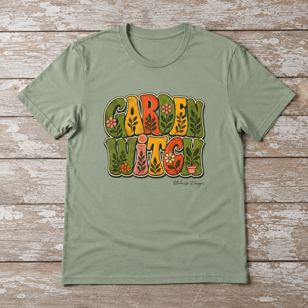 Flat lay of a Sage Garden Witch T-shirt featuring retro botanical lettering in olive, mustard, orange, and pink with flowers and leafy details.