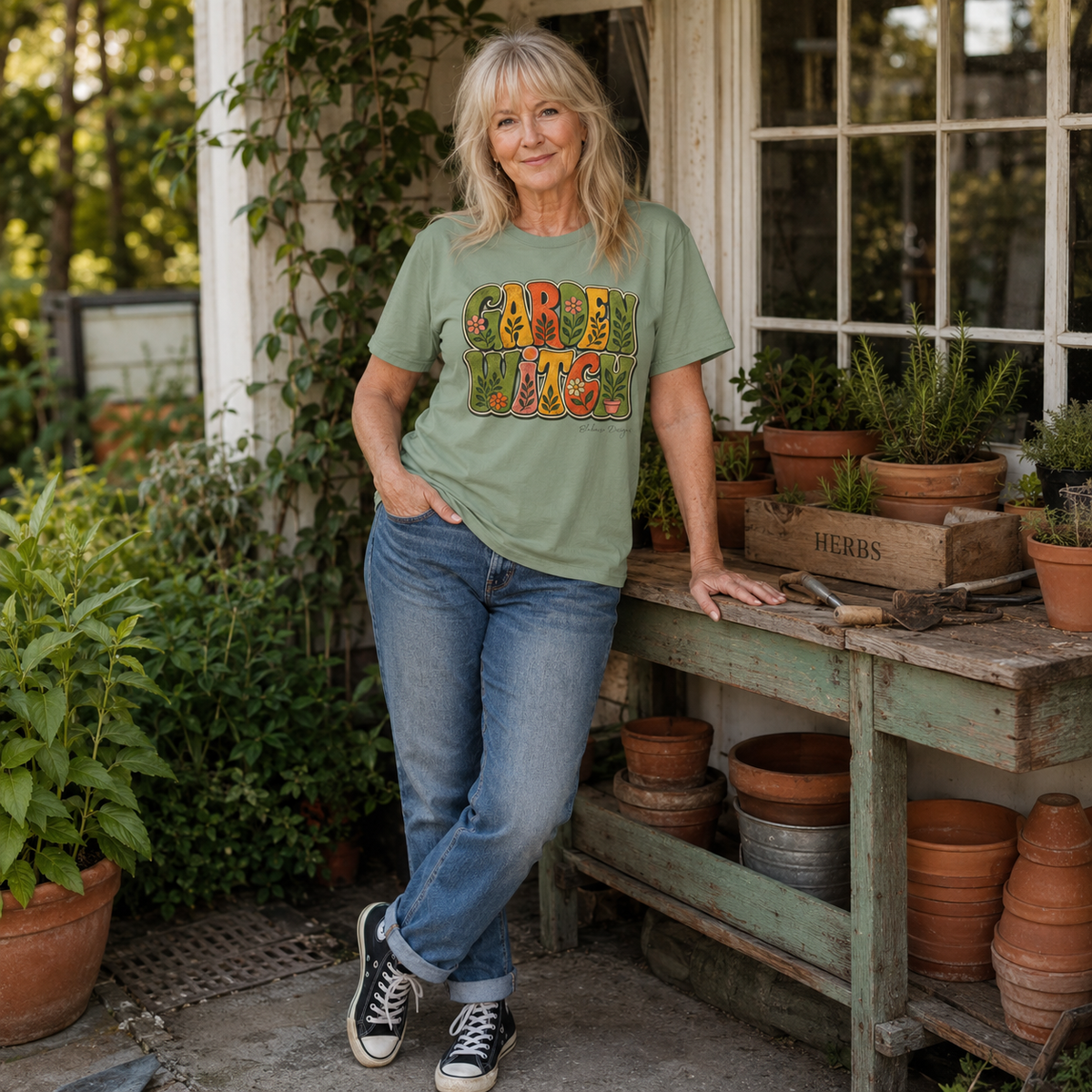 Lifestyle image of a woman wearing a Sage Garden Witch T-shirt beside a weathered potting bench with terracotta pots, herbs, and garden tools.