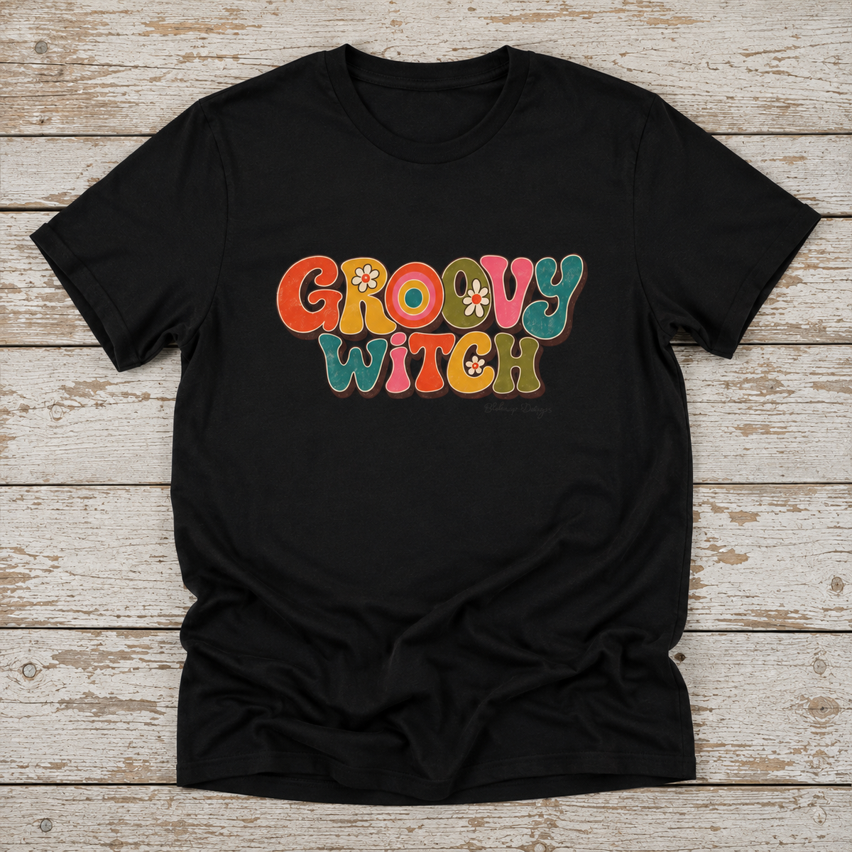 Flat lay of a Groovy Witch T-shirt featuring retro 70s inspired lettering in warm earthy colors with floral details and distressed texture.