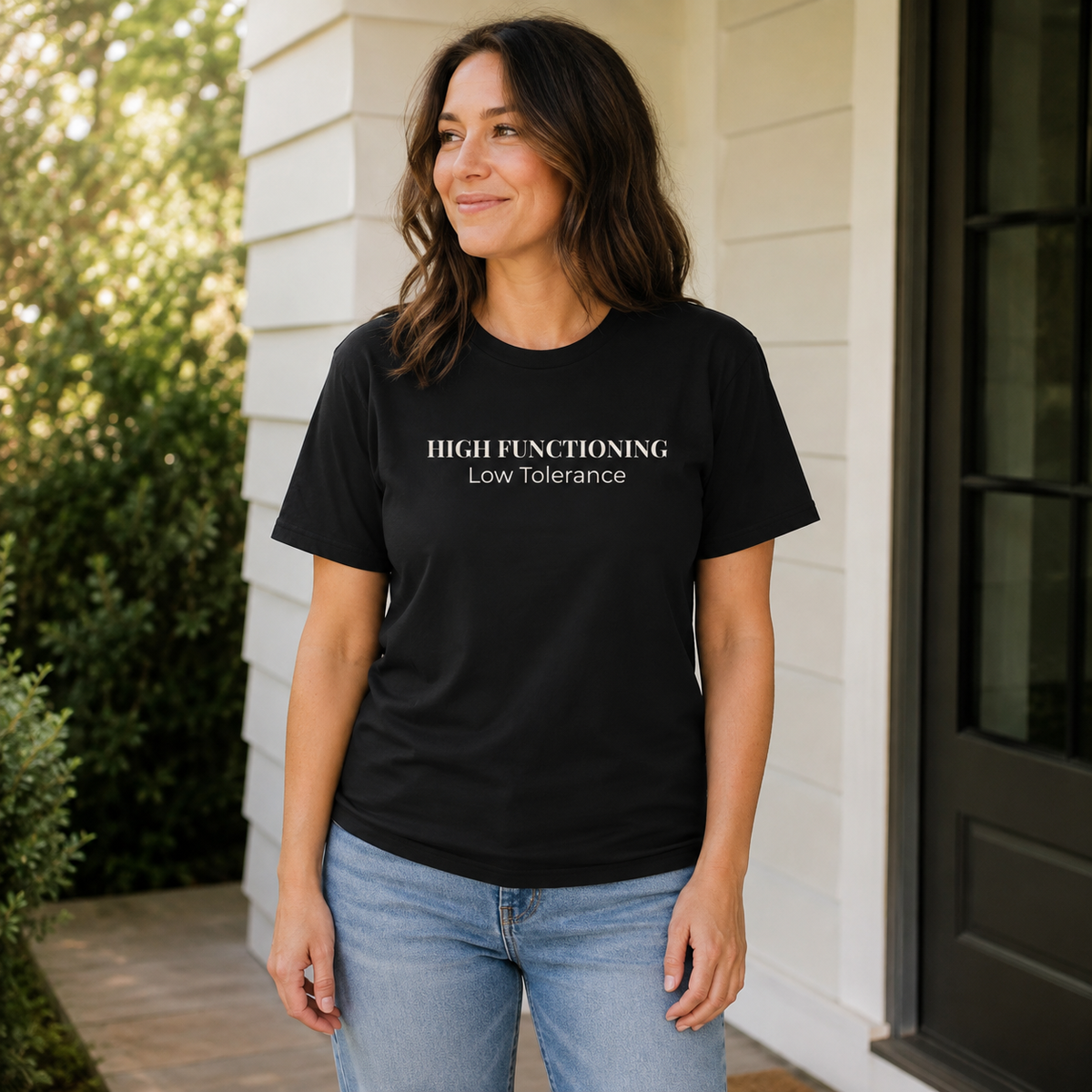 Woman wearing a black High Functioning Low Tolerance unisex tee while standing casually on a porch.