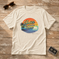 Natural unisex graphic tee featuring “Josie’s on a Vacation Far Away” over a retro tropical sunset with palm trees and ocean waves.