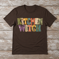 Dark Chocolate Kitchen Witch T-shirt displayed on a weathered wood surface with colorful scrapbook style lettering, herbs, recipe notes, gingham, and kitchen details.