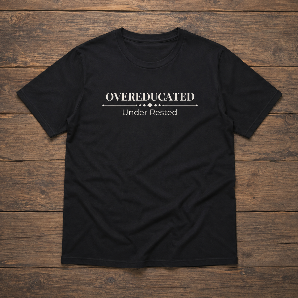 Black unisex tee laid flat on a warm wood surface featuring the Overeducated Under Rested design across the chest.