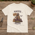 Women's or unisex Gildan Softstyle graphic tee featuring Diana the raccoon holding a Coexist sign beneath the phrase "Peaceful Does Not Mean Passive."