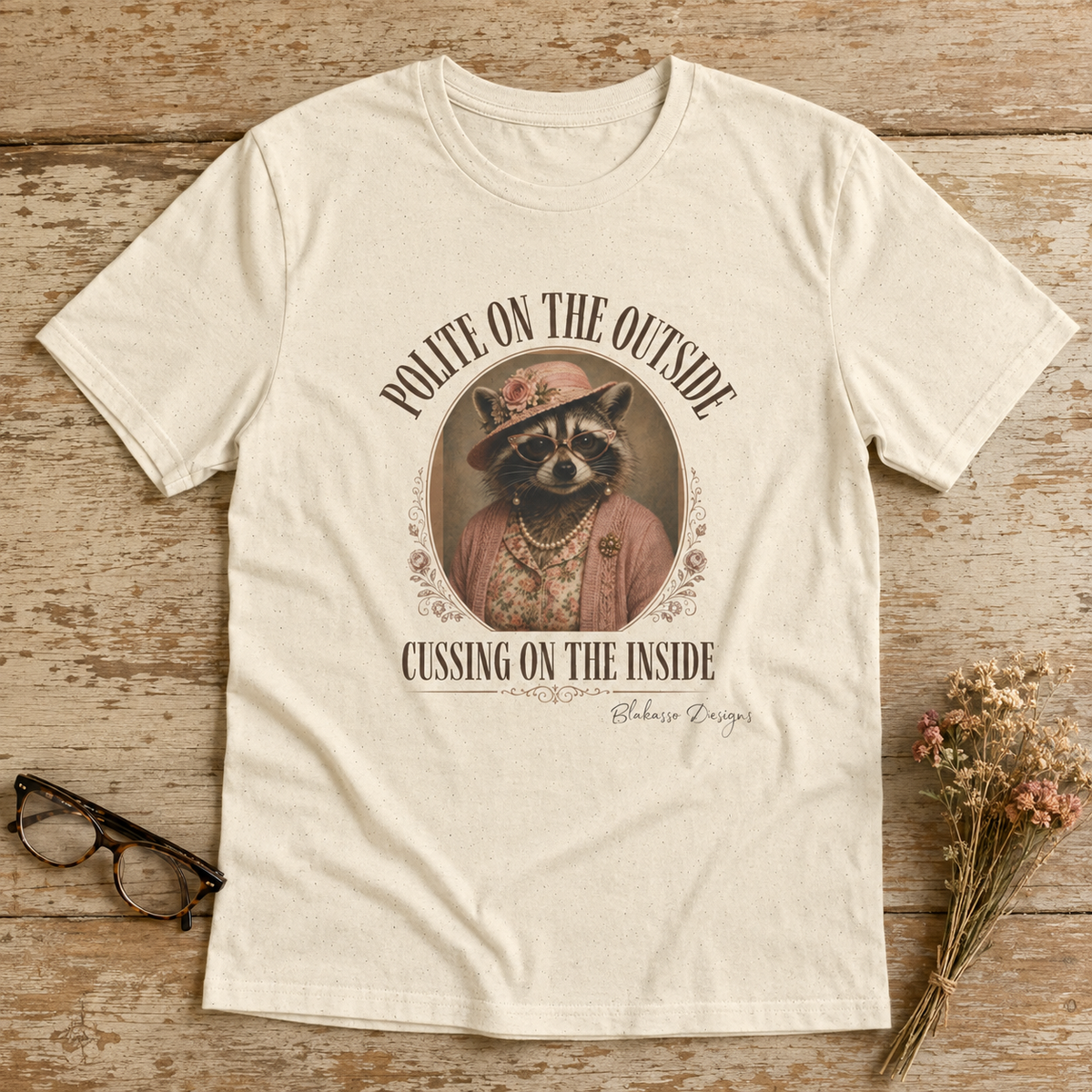 Natural unisex tee featuring Miss Cordelia the raccoon in pearls and a pink church hat with the words “Polite on the Outside, Cussing on the Inside.”