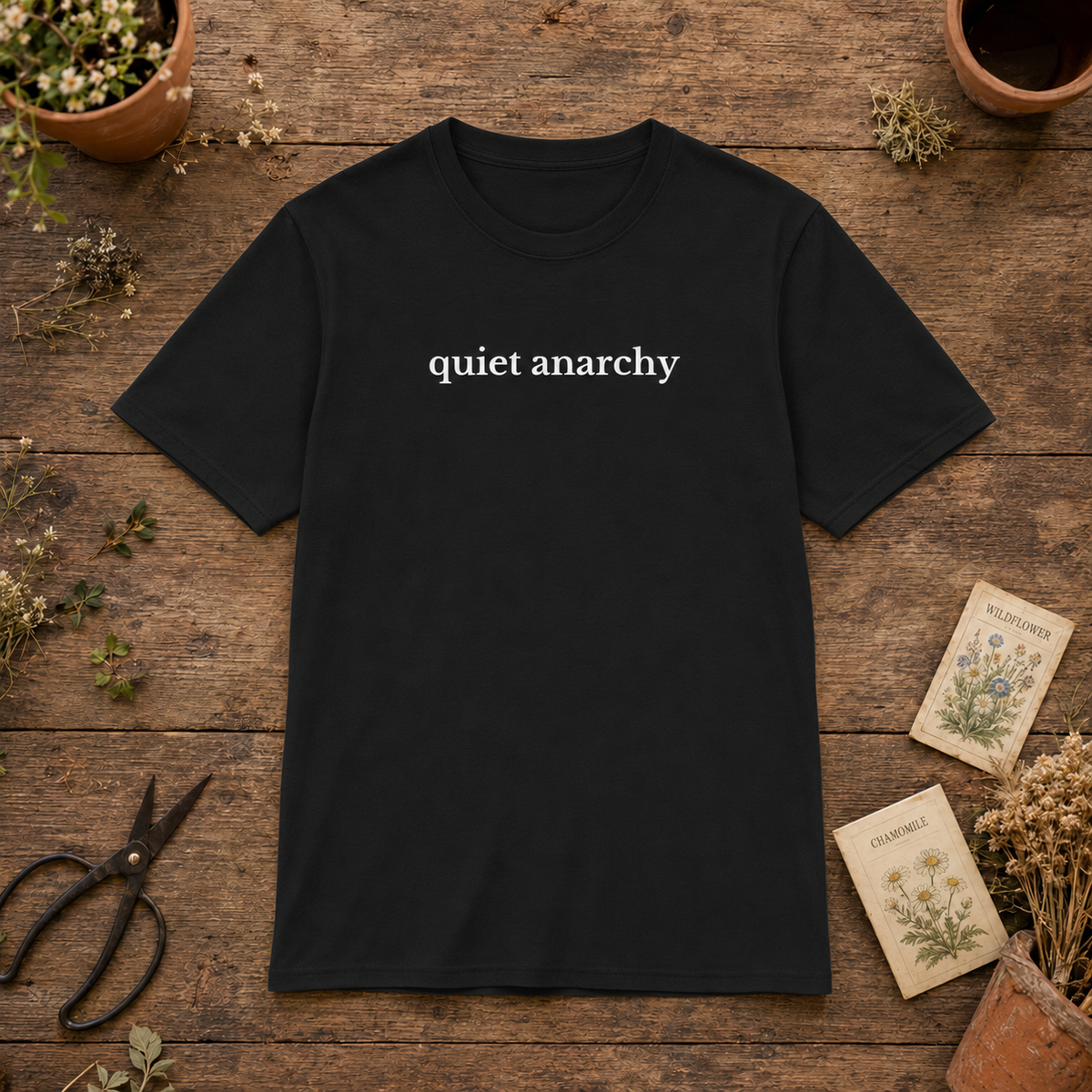 A black short-sleeve t-shirt with the words 'quiet anarchy' printed in white serif text across the chest, laid flat on a rustic wooden surface surrounded by dried flowers, scissors, and small potted plants.