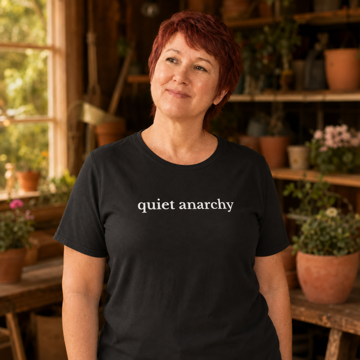 A woman with short red hair wearing a black t-shirt with the words 'quiet anarchy' printed in white lowercase letters across the chest, standing in a warm, sunlit greenhouse or garden center filled with potted plants.