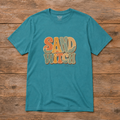 Flat lay of a Heather Galapagos Blue Sand Witch T-shirt featuring retro lettering in orange, mustard, cream, and seafoam with sun and wave details.
