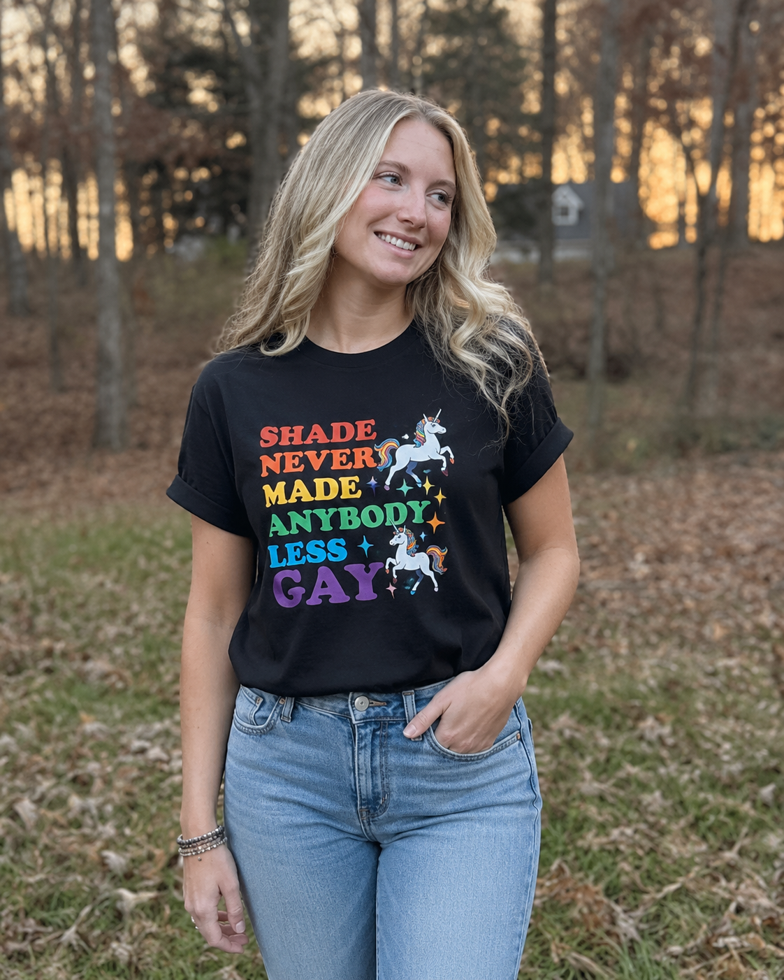 Woman wearing a black Shade Never Made Anybody Less Gay graphic tee featuring rainbow lettering and colorful unicorns.