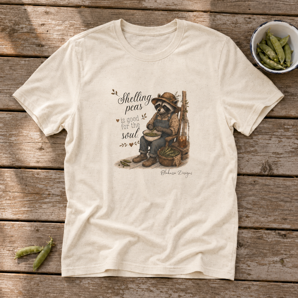 Natural unisex graphic tee featuring Miss Sammye the raccoon shelling peas with the words “Shelling Peas Is Good for the Soul.”
