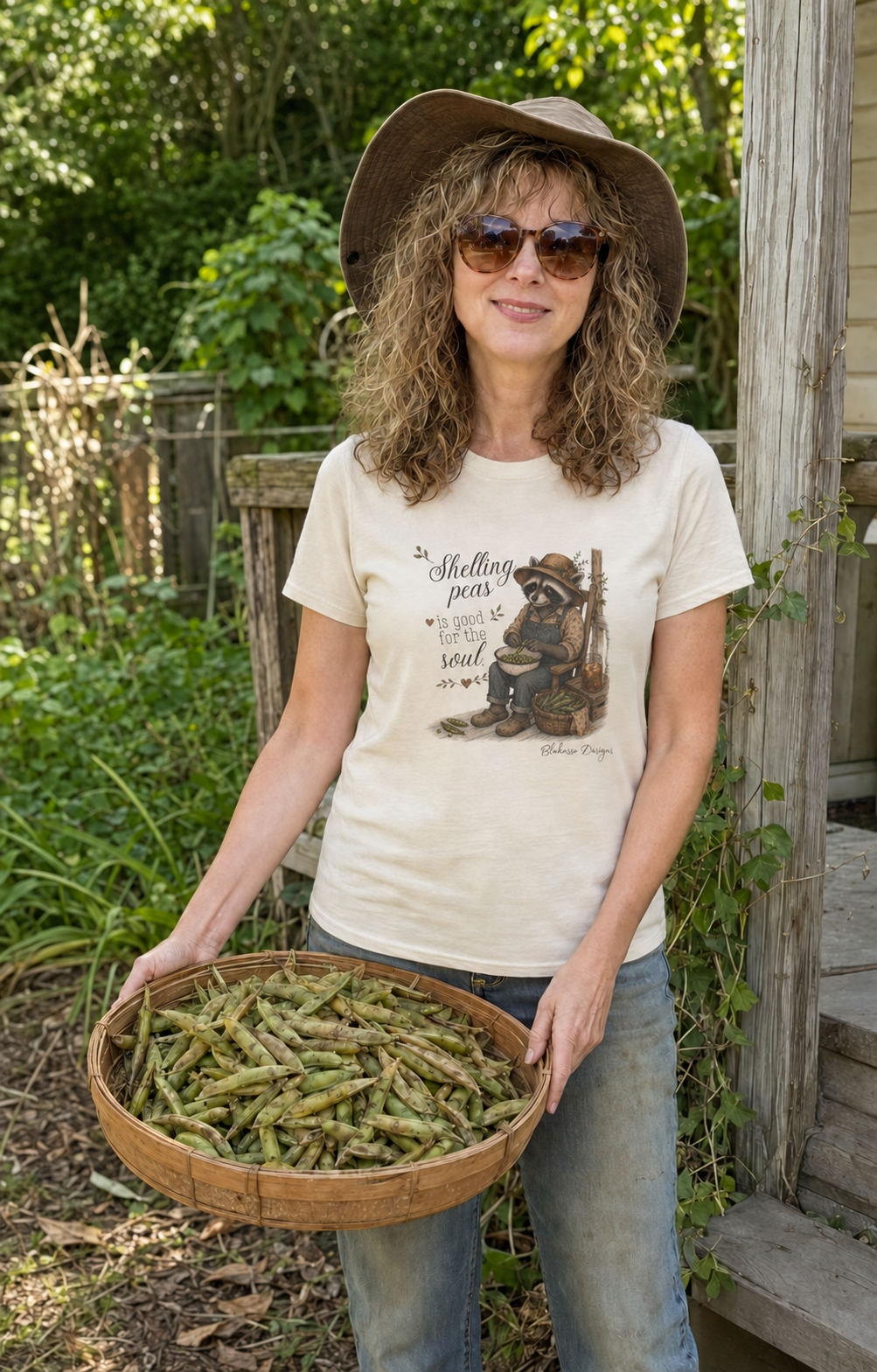 Woman wearing Shelling Peas Is Good for the Soul raccoon shirt while holding a basket of peas