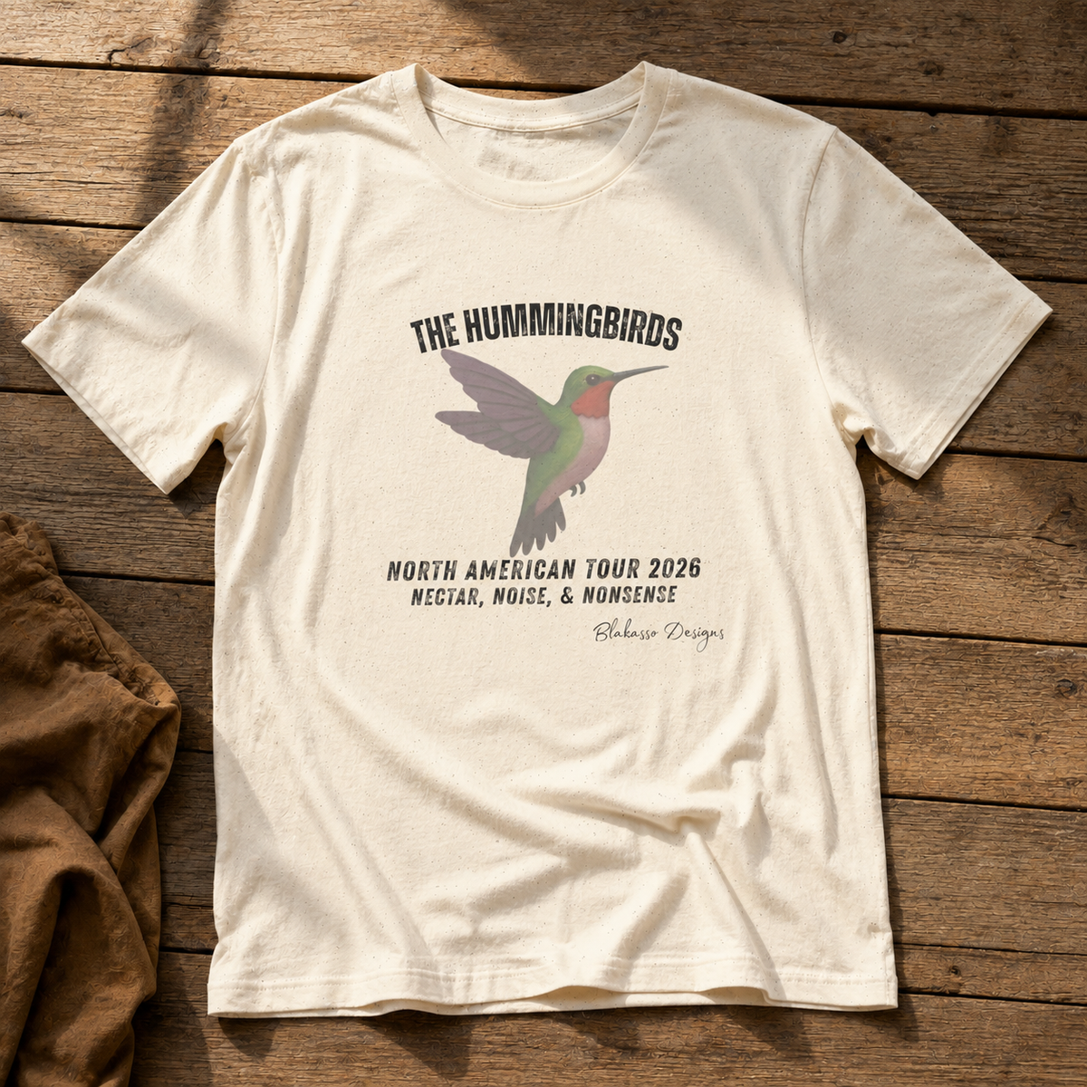 An off-white graphic t-shirt laid flat on a rustic wooden background. The shirt features a printed design of a hummingbird with the text 'THE HUMMINGBIRDS NORTH AMERICAN TOUR 2026 NECTAR, NOISE, & NONSENSE' and 'Blakasso Designs' written below.