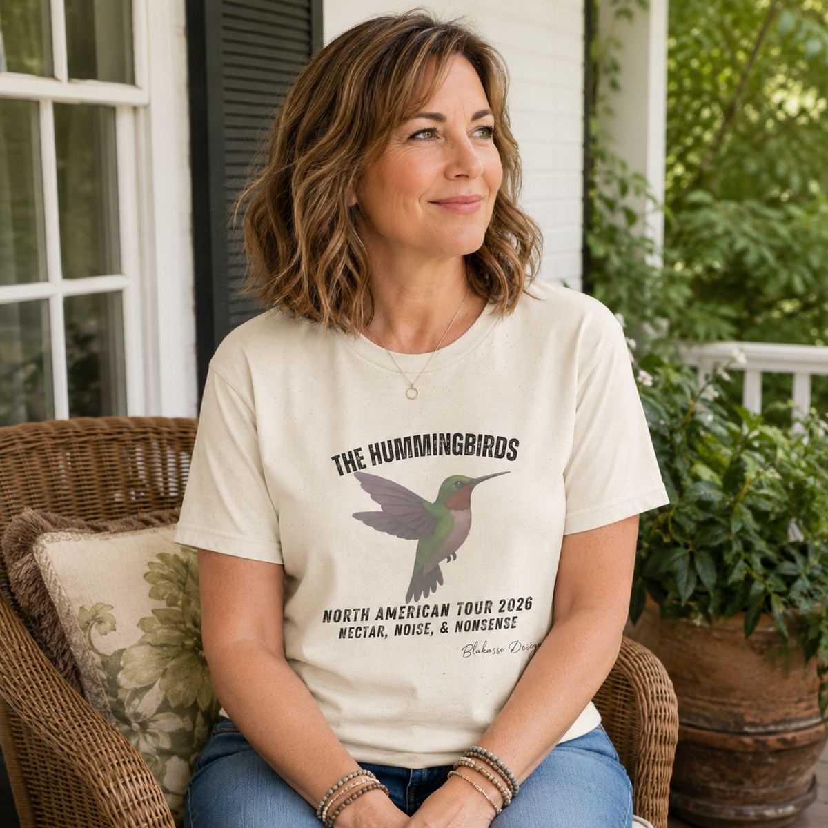 Middle aged woman wearing a natural The Hummingbirds North American Tour 2026 graphic T-shirt while sitting on a porch.