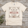 Flat lay of a Gildan Softstyle T-shirt featuring The Older I Get, the Heller the Naw design in a cross stitch inspired style with flowers, a decorative border, and a cat at the bottom.