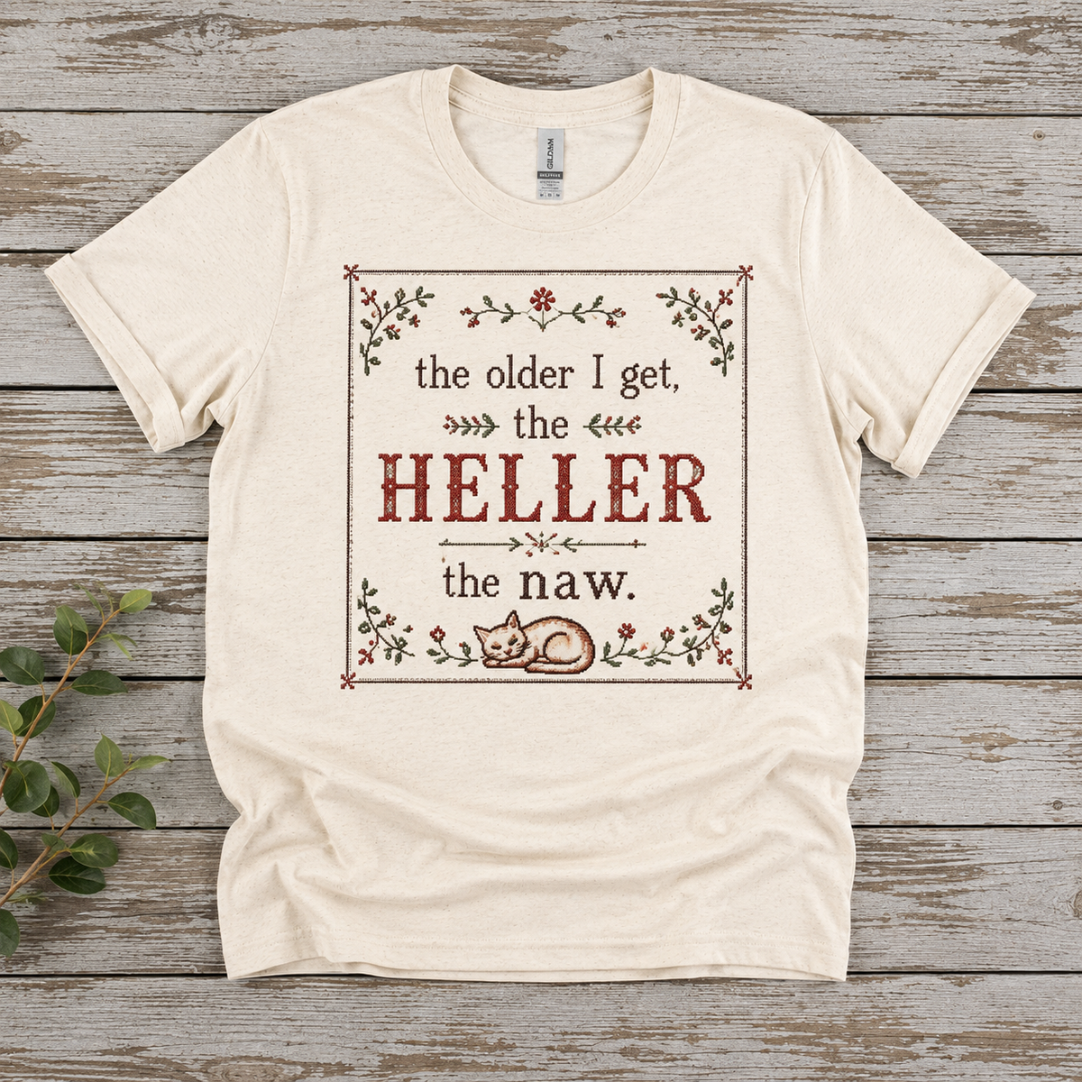 Flat lay of a Gildan Softstyle T-shirt featuring The Older I Get, the Heller the Naw design in a cross stitch inspired style with flowers, a decorative border, and a cat at the bottom.