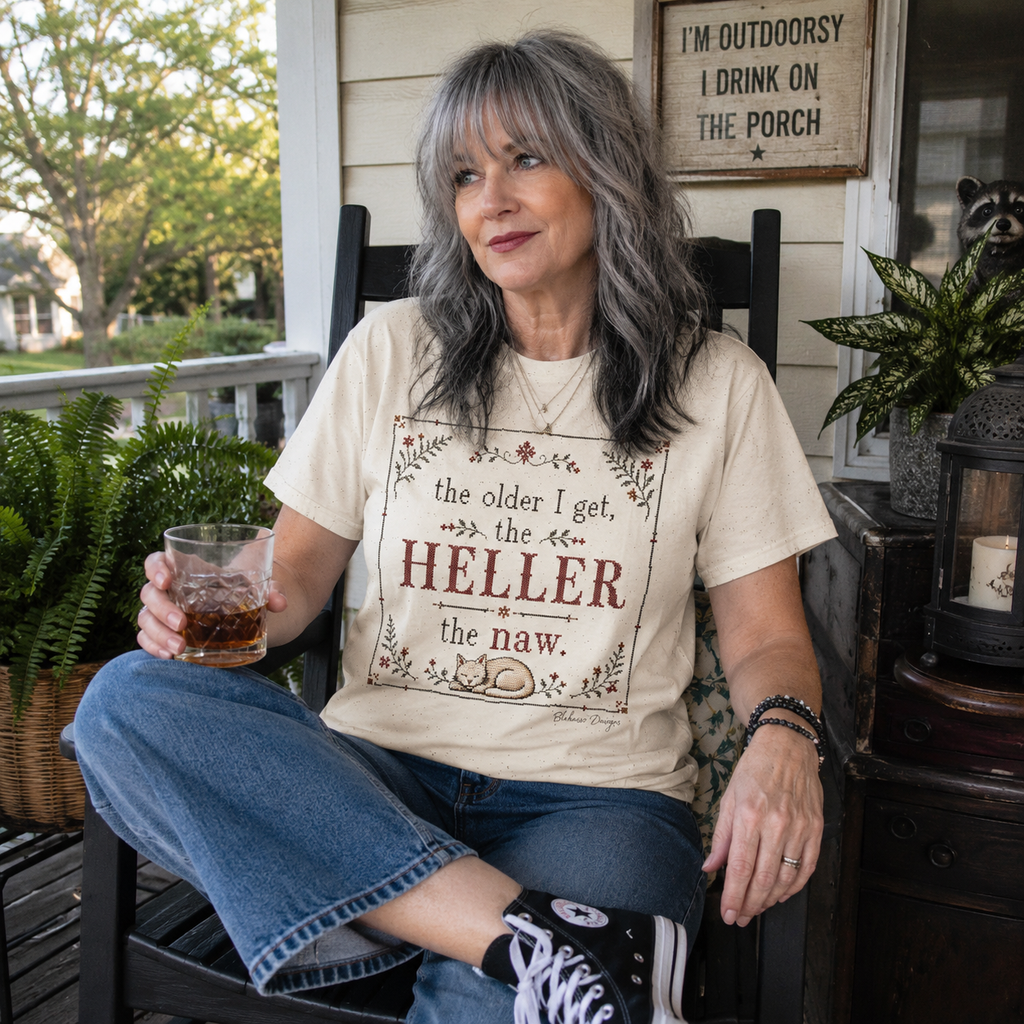 A woman with gray hair sits on a porch chair wearing a cream graphic t-shirt with a brown and red text print that reads 'the older I get, the HELLER the naw.' She is holding a glass and wearing blue jeans and black sneakers.